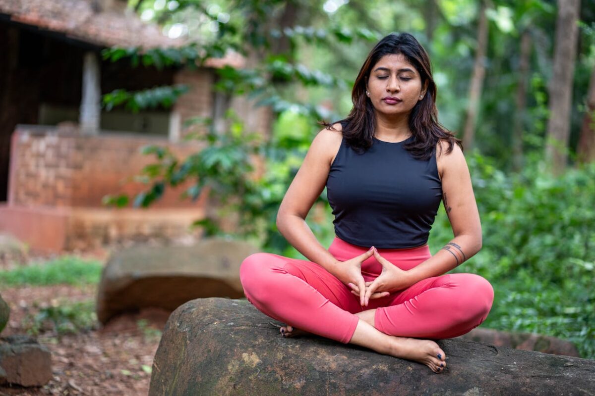 A woman practicing meditation outdoors on a rock, surrounded by lush greenery, promoting tranquility and mindfulness.