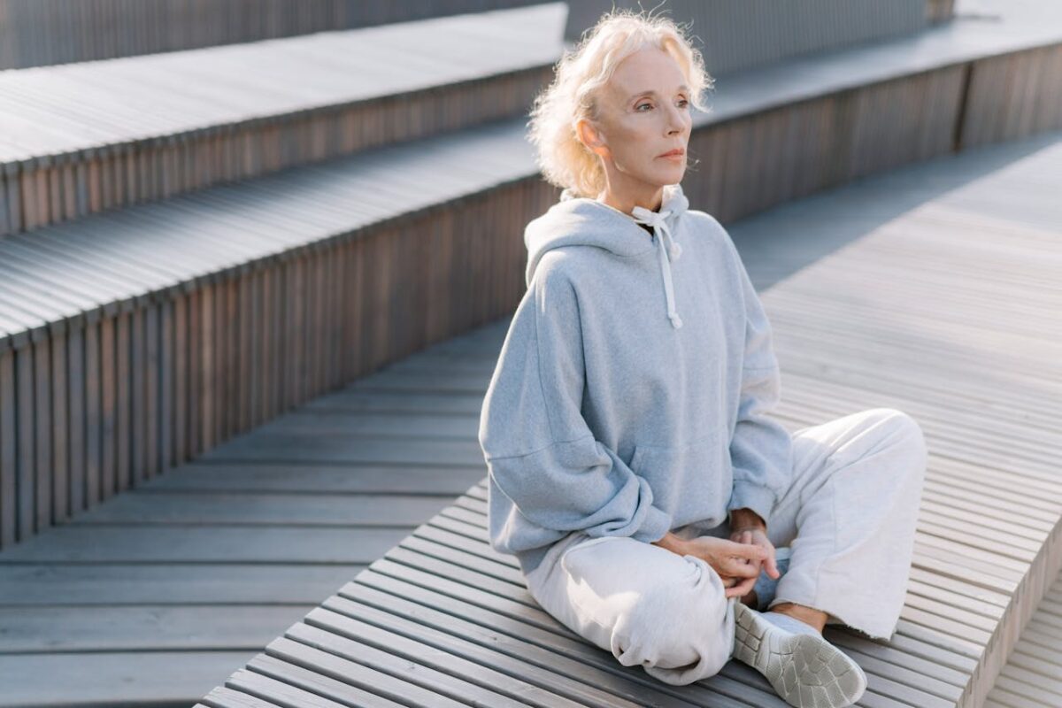 Elderly woman meditating outdoors in sunlight. Relaxation and healthy lifestyle in soft gray sportswear.