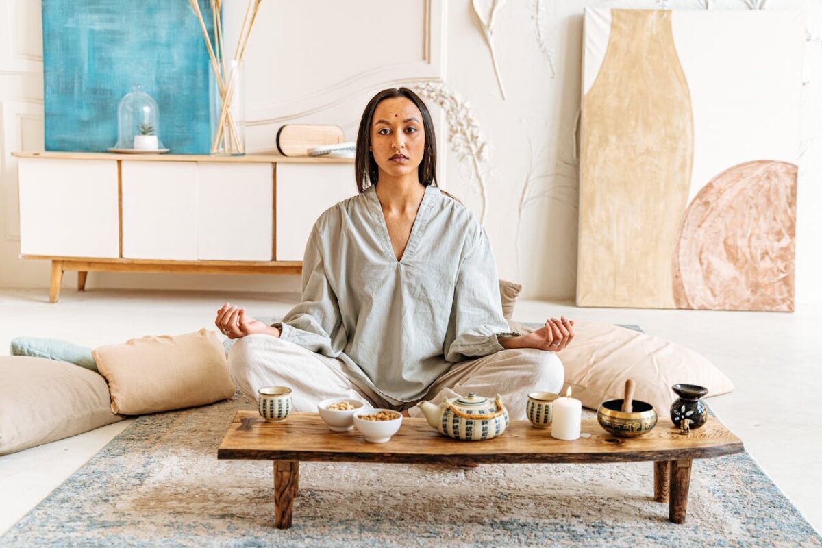Woman peacefully meditating indoors surrounded by cushions and tea set, promoting tranquility.